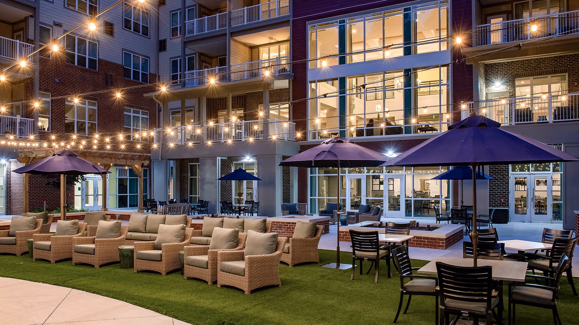 Outdoor courtyard area of a senior living facility at night with string lights overhead, multiple wicker armchairs with cushions arranged in rows, several tables with chairs and large purple umbrellas, and a multi-story building with large windows in the background.