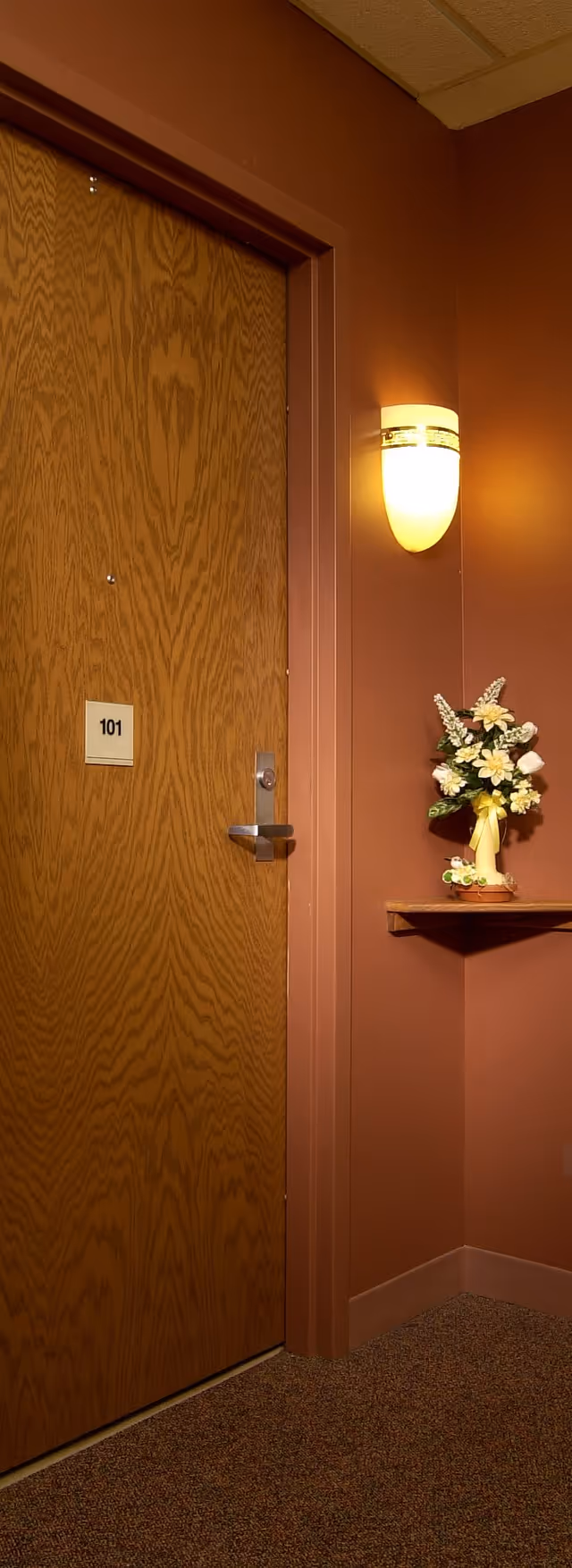 A wooden door labeled with the number 101 in a hallway with brown walls and carpeted floor. To the right of the door, there is a wall-mounted light fixture emitting warm light and a small shelf holding a vase with artificial flowers.