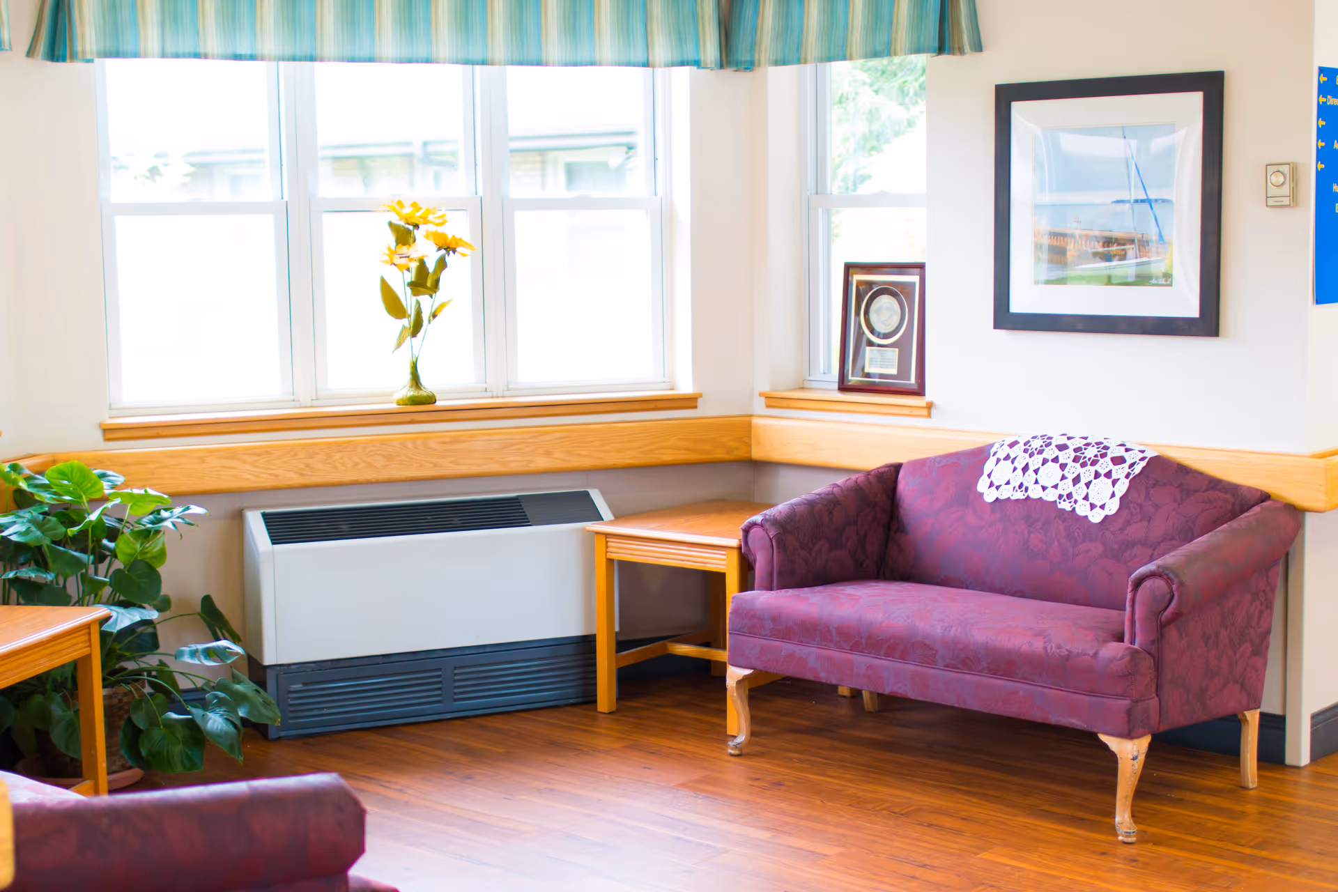 A cozy corner of a living room with a purple upholstered loveseat adorned with a white crocheted doily on the backrest. Next to the loveseat is a wooden side table, and a heating unit is positioned beneath a set of three windows with a striped valance. A vase with yellow flowers sits on the windowsill, and a green leafy plant is placed on the floor beside the table. A framed picture and a plaque are displayed on the window ledge, and the room has wooden flooring.