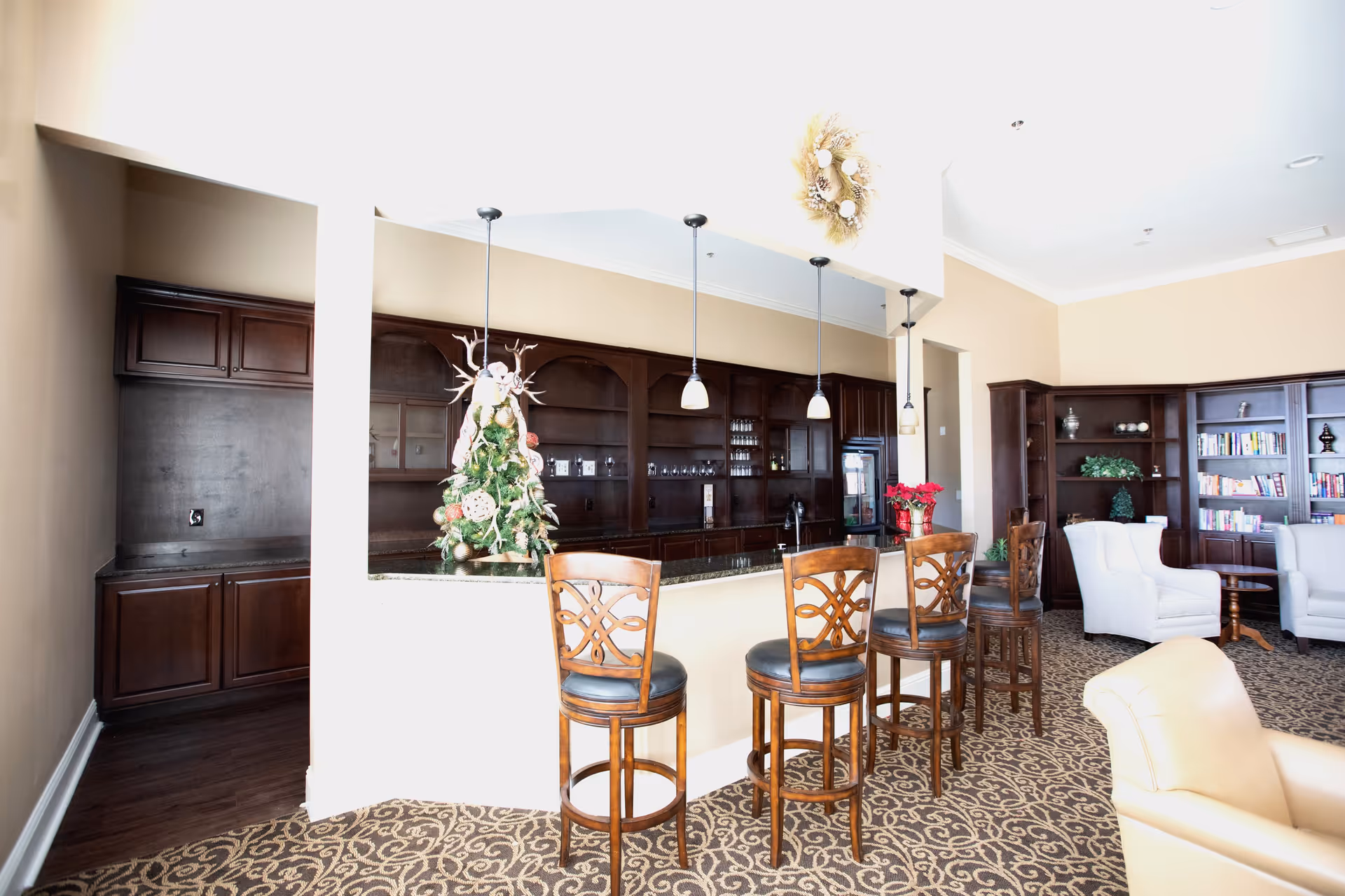 A cozy common area with a bar counter featuring four wooden bar stools with cushioned seats. Behind the bar are dark wooden cabinets and shelves with glassware and a small refrigerator. The room is decorated with a small Christmas tree on the counter and a wreath hanging above. To the right, there are bookshelves filled with books and two white armchairs around a small wooden table. The floor is carpeted with a patterned design.