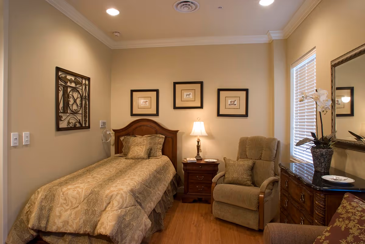 A cozy bedroom with a single bed covered in a patterned beige and green comforter, a wooden headboard, and matching pillows. Next to the bed is a wooden nightstand with a lamp and some items on it. There is a comfortable beige recliner chair with a matching pillow beside the nightstand. On the right side, there is a wooden dresser with a black top, a decorative vase with white flowers, and a large mirror above it. The walls are painted light beige and decorated with three framed pictures above the bed and a decorative metal wall art on the left wall. A window with white blinds is on the right side, letting in natural light. The floor is wooden.