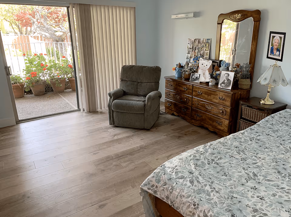 A cozy bedroom with a floral bedspread, a wooden dresser with a mirror, a cushioned armchair, and a side table with a lamp. There is a sliding glass door with vertical blinds leading to an outdoor patio with potted plants and flowers.