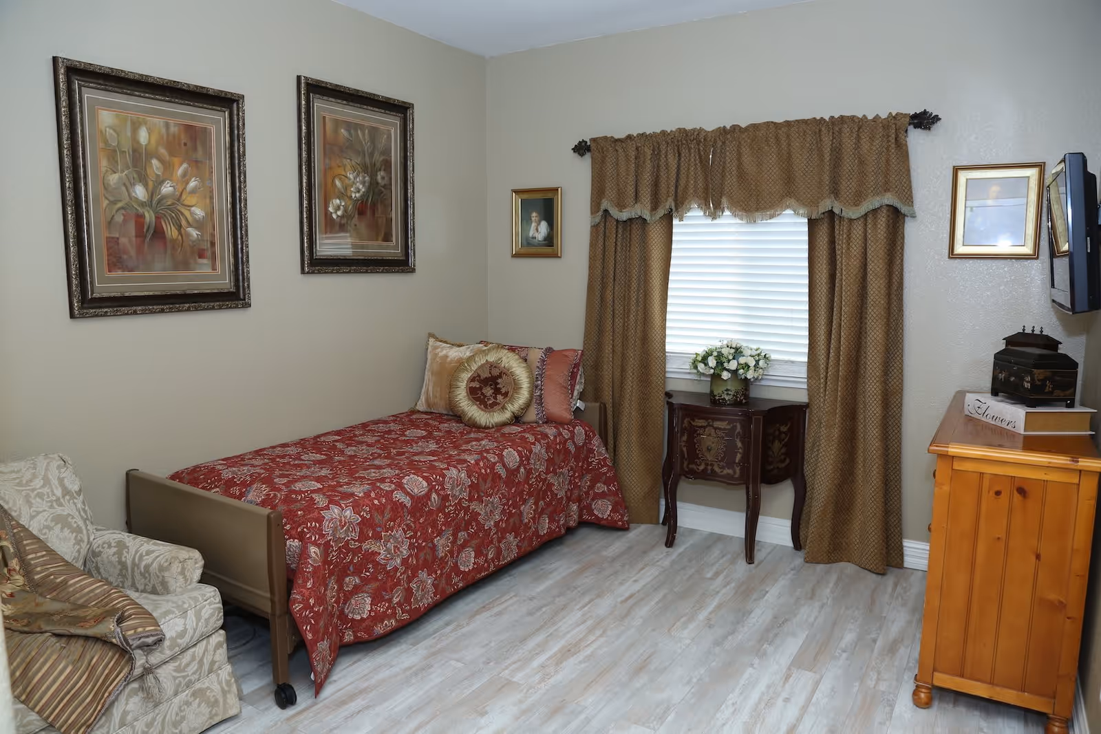 A cozy bedroom with a single bed covered in a red floral bedspread and decorative pillows. There is a beige armchair with a striped cushion next to the bed. The walls are adorned with framed floral artwork and a small portrait. A window with brown curtains and a valance is centered on the far wall, with a small ornate table holding a vase of white flowers beneath it. A wooden dresser with a small sign and decorative boxes is on the right side, and a flat-screen TV is mounted on the wall above it. The floor has light-colored wood laminate.