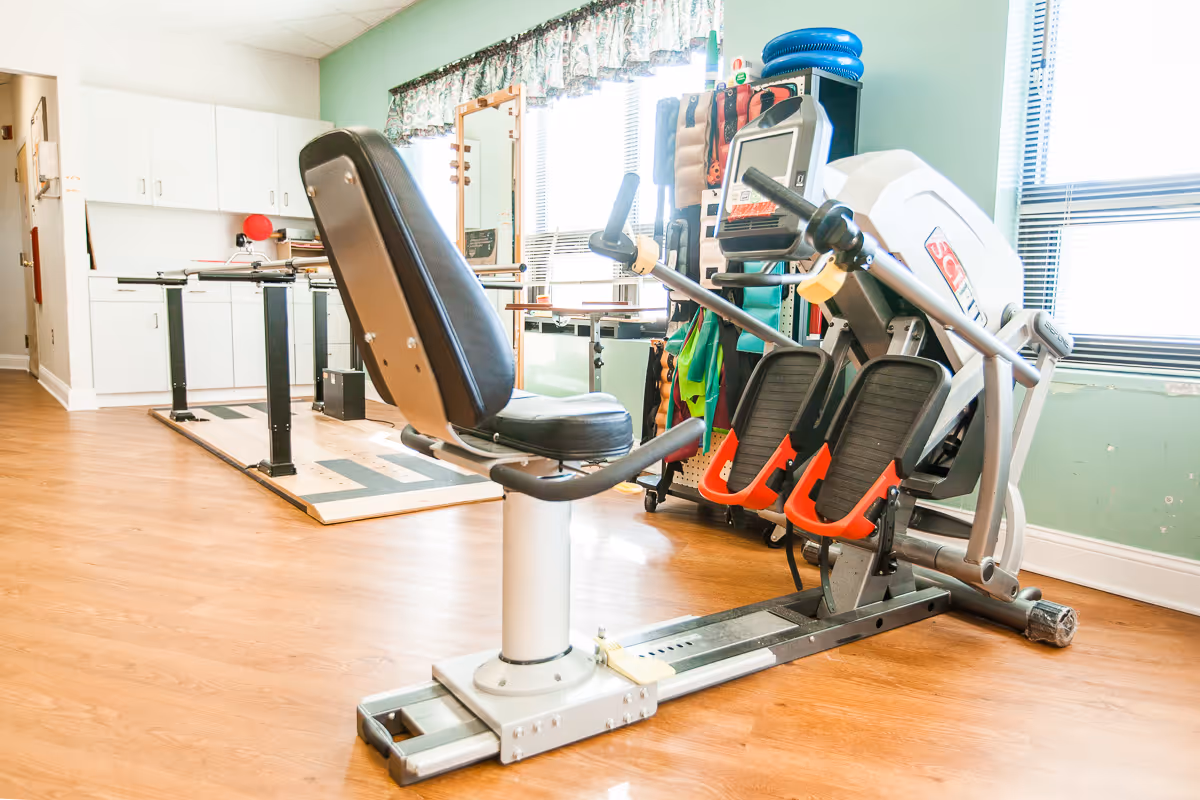 A bright room with exercise equipment including a seated rowing machine and a parallel bar setup. The room has wooden flooring, light green walls, and windows with blinds and floral curtains letting in natural light. There are cabinets and a mirror in the background along with various exercise accessories.