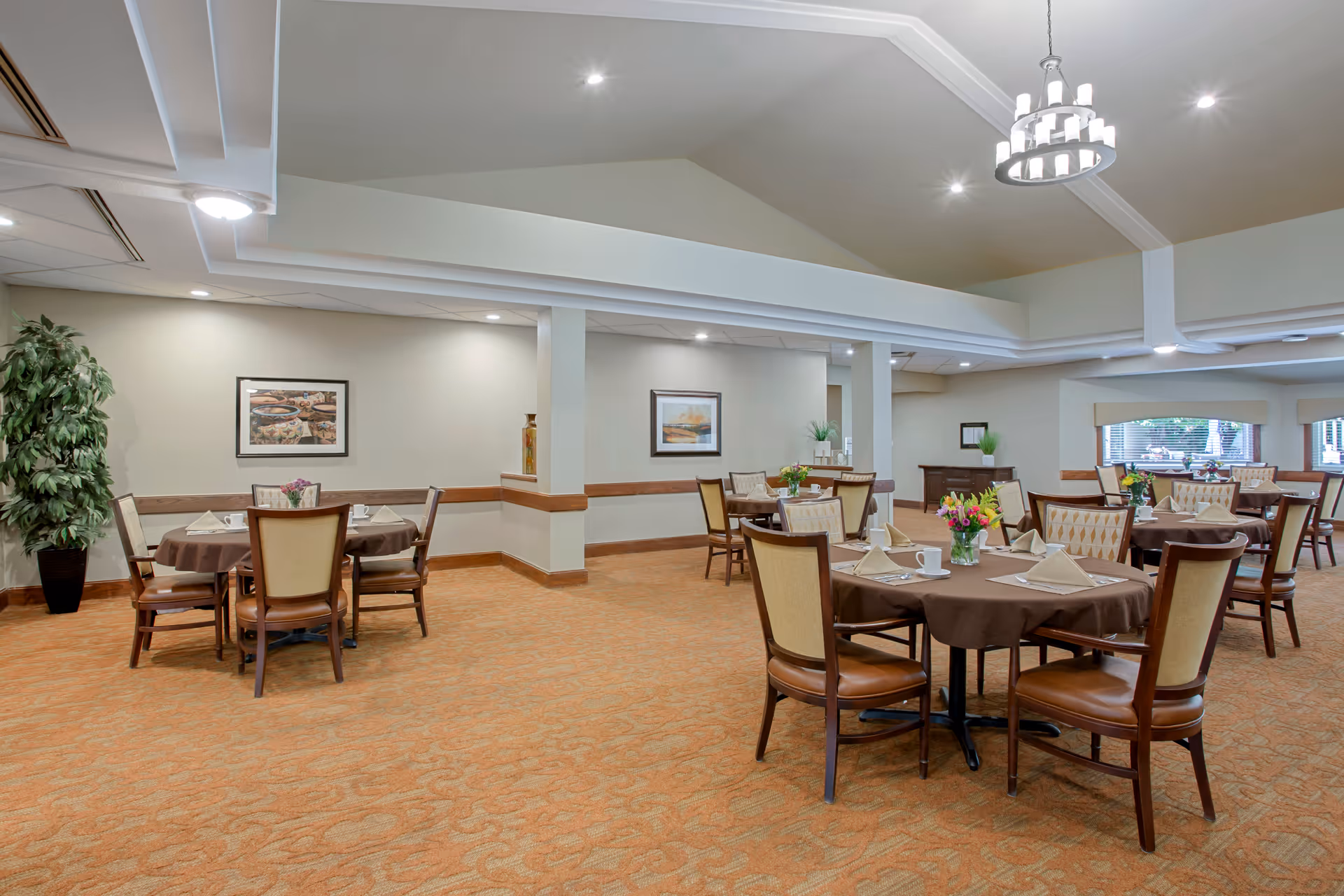 A spacious dining room with several round tables covered with brown tablecloths, each set with napkins, cups, and small flower arrangements. The room has beige walls, carpeted floors, framed artwork, and large windows letting in natural light. There is a chandelier hanging from the ceiling and a potted plant in one corner.