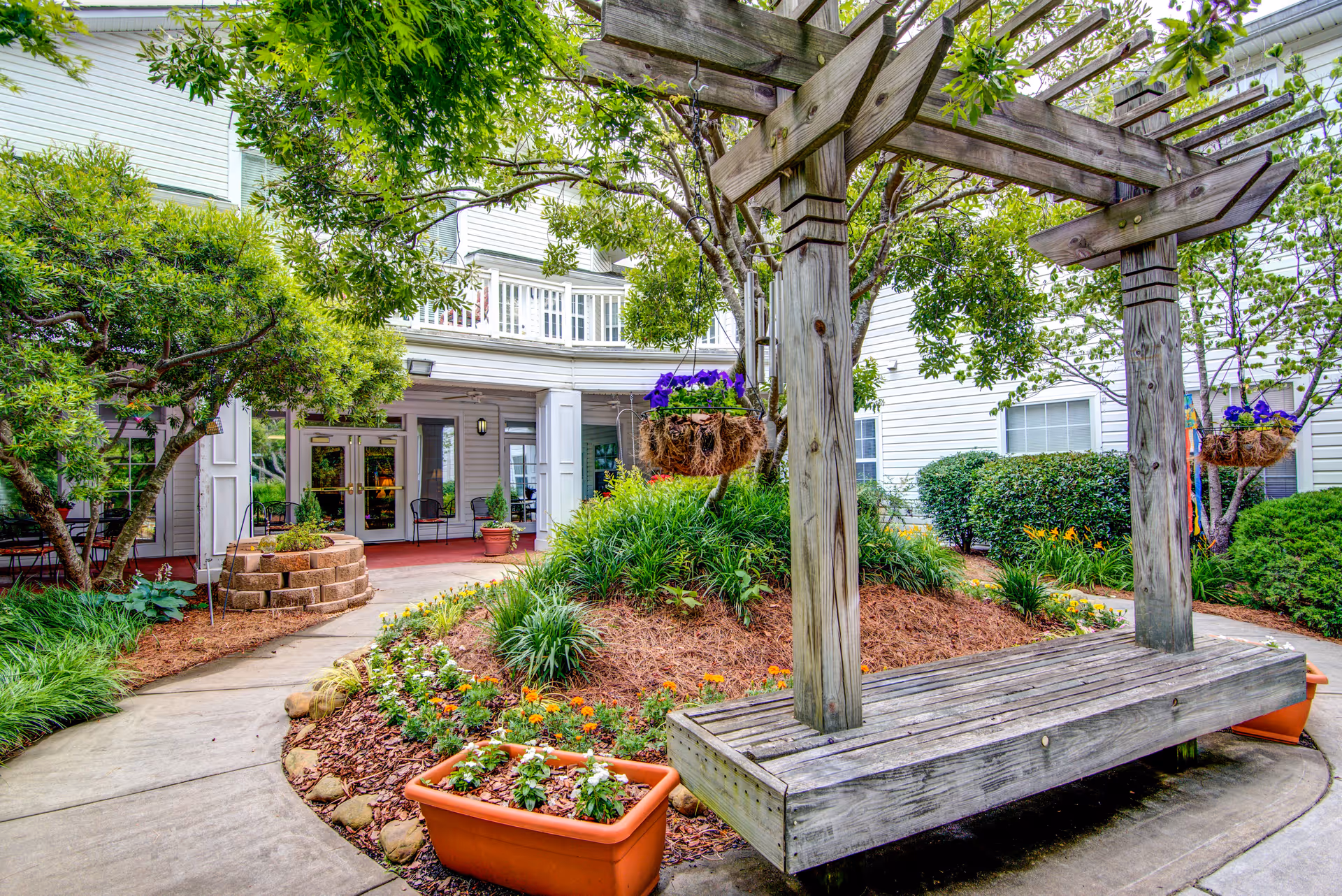 Outdoor garden area at Oaks at Snellville featuring a wooden pergola with hanging flower baskets, a wooden bench, various plants and flowers, and a pathway leading to the entrance of a white building with large windows and doors.