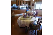 Interior view of a dining area with round tables covered with white tablecloths, set with plates, cups, and floral centerpieces. Wooden chairs surround the tables, and a kitchen area with cabinets and appliances is visible in the background.