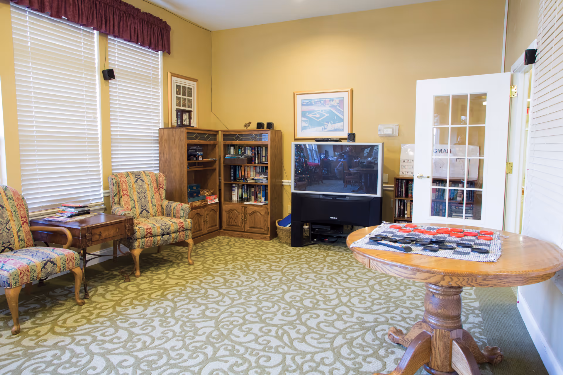 A cozy common room with patterned carpet and yellow walls. There are two upholstered armchairs with floral patterns next to a wooden side table. A wooden bookshelf filled with books is against the wall. A large flat-screen TV sits on a black stand. A round wooden table with a checkers game set up is in the foreground. The room has two large windows with white blinds and a white door with glass panes.