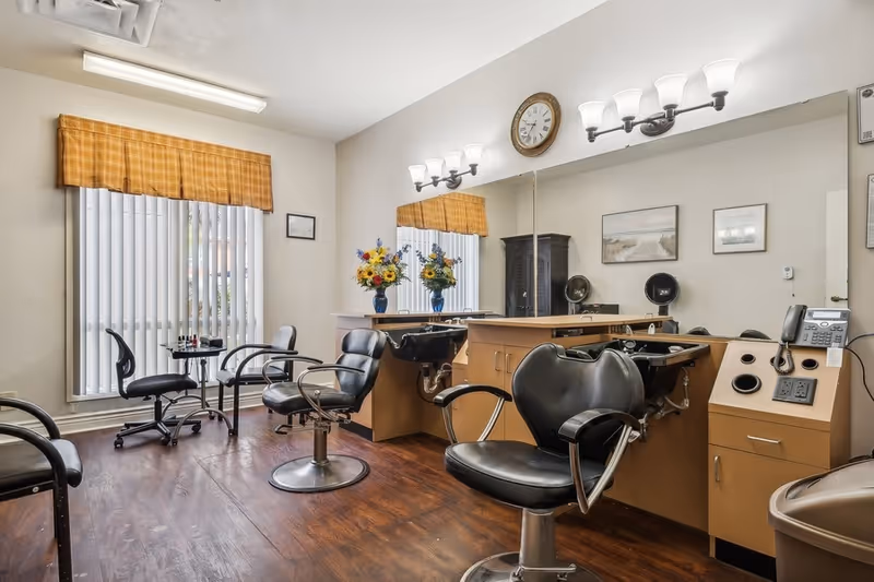 Interior of a salon area with black salon chairs, hair washing sinks, a large mirror with wall-mounted lights above, a clock, and a window with vertical blinds and a yellow valance. There are flowers in a vase on the counter and framed pictures on the wall.