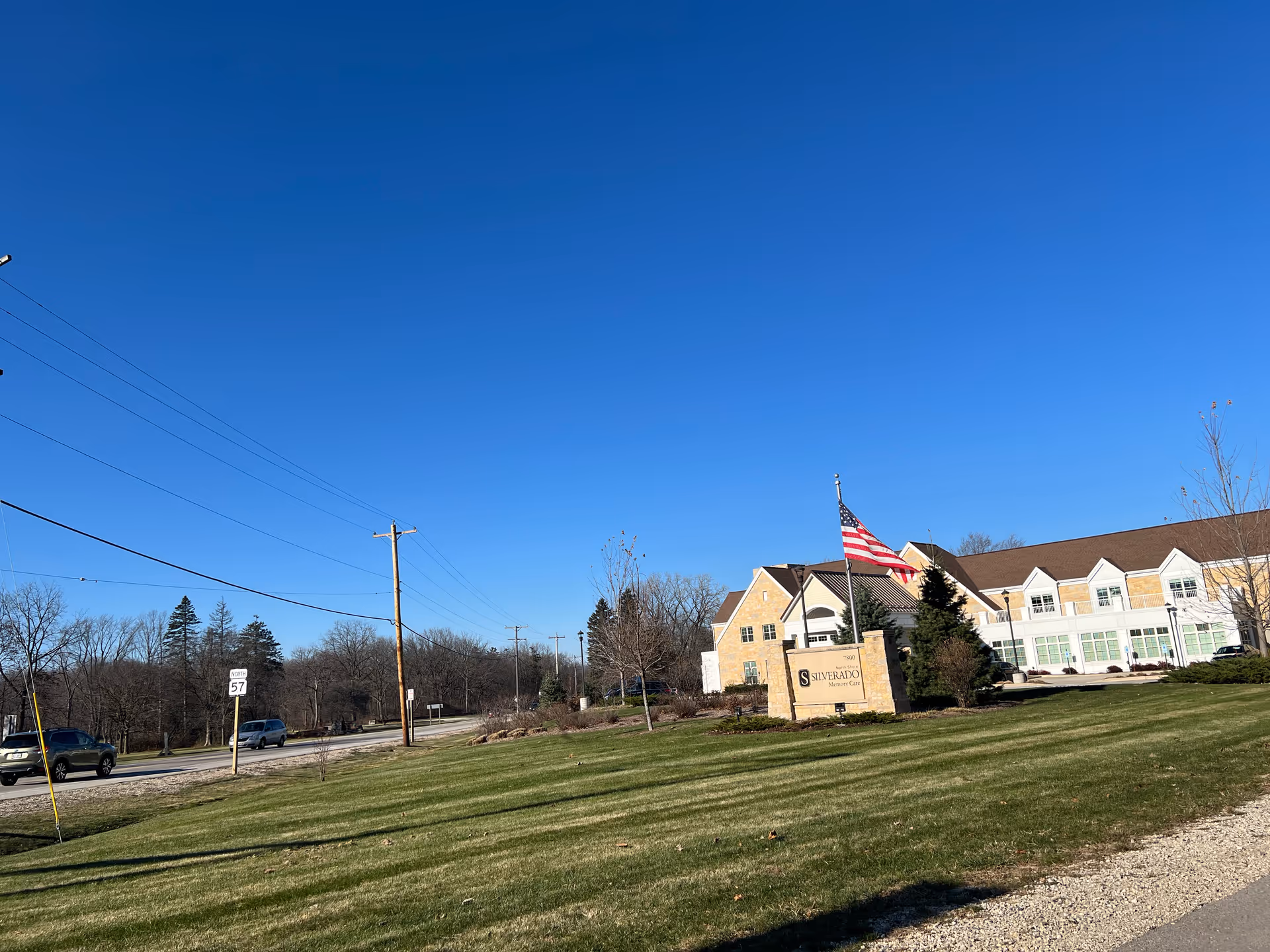Front exterior of the Silverado North Shore Memory Care building with its sign and American flag on a lawn beside a road under a clear blue sky.