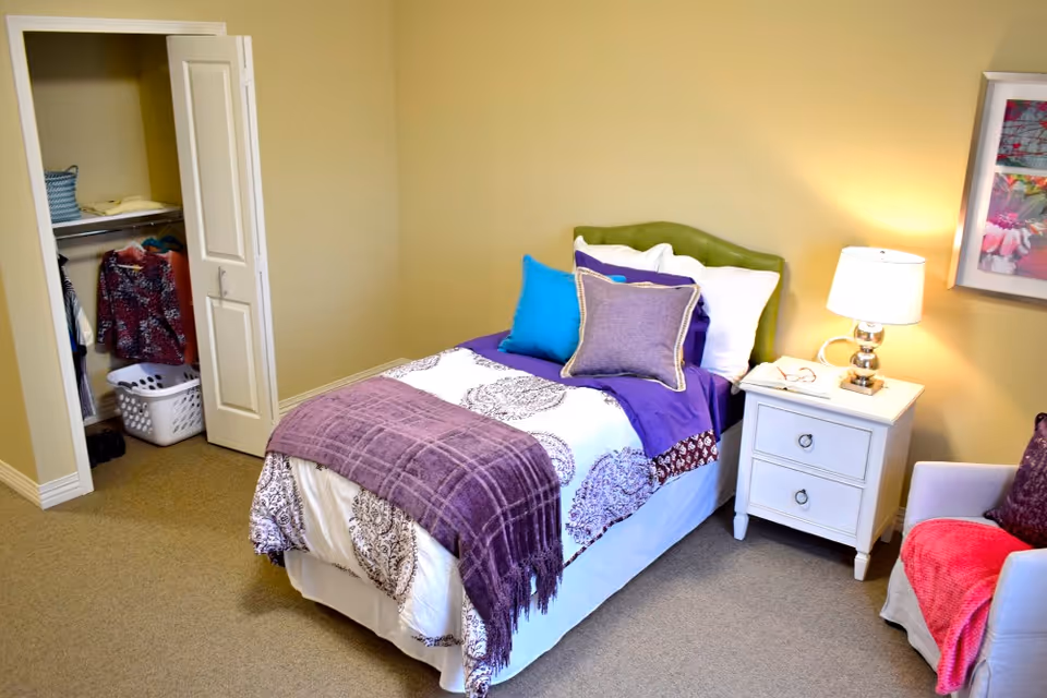 A cozy bedroom with a single bed dressed in white and purple bedding, multiple pillows, and a purple throw blanket. Next to the bed is a white nightstand with a lamp and a pair of glasses. An armchair with a purple pillow and a pink throw is positioned nearby. An open closet with clothes hanging and a laundry basket is visible in the background. The walls are painted light yellow and there is a framed floral artwork on the wall.