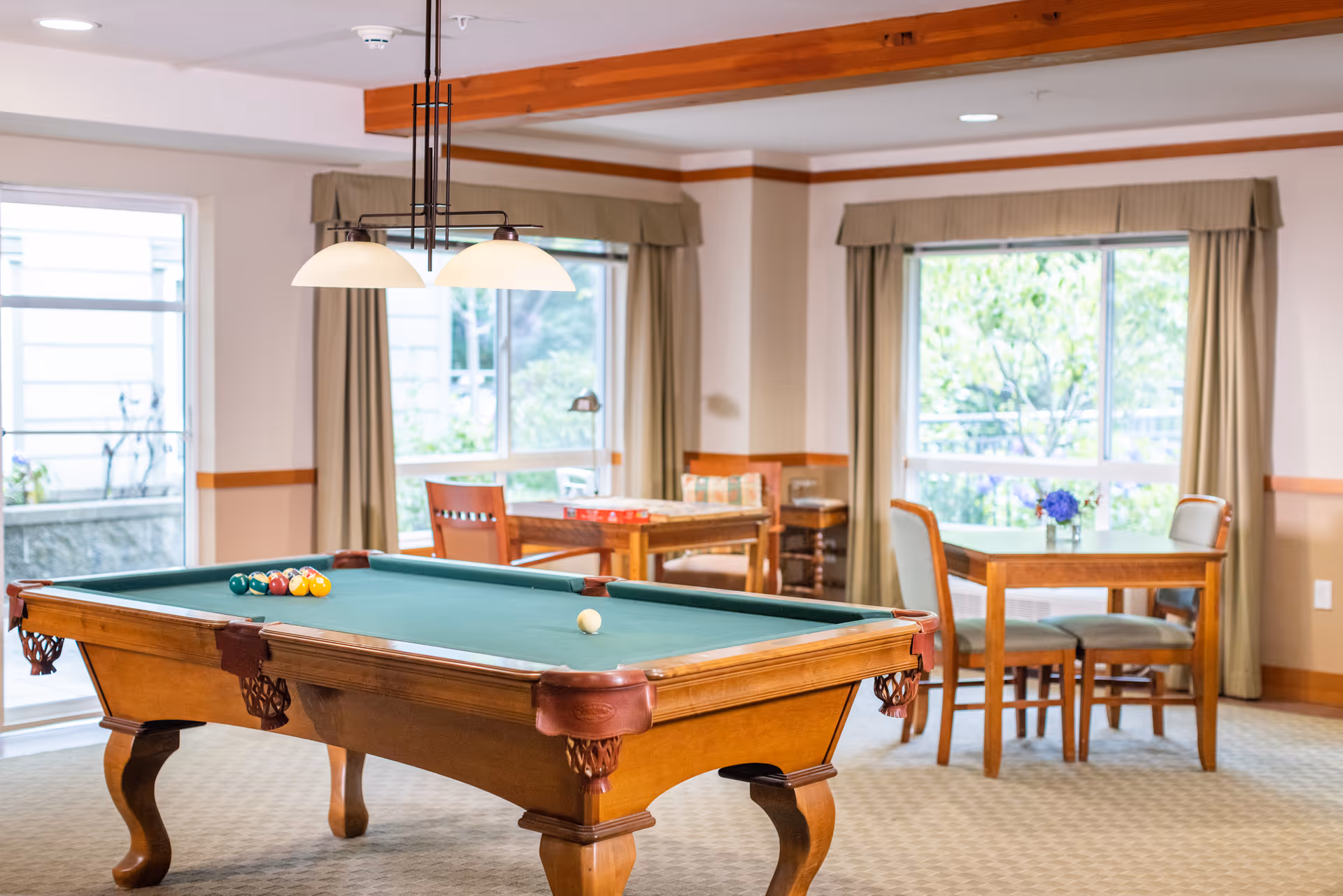 A well-lit recreational room with a wooden pool table in the foreground, set up with billiard balls and a cue ball. In the background, there are two wooden tables with chairs, one with a vase of purple flowers. Large windows with beige curtains allow natural light to fill the room.