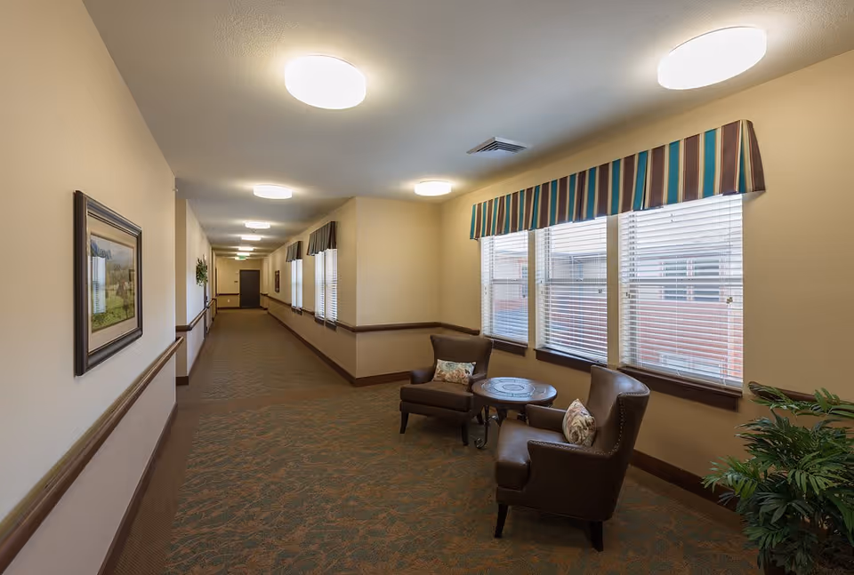 A long, well-lit hallway in a senior living facility with beige walls and carpeted floor. On the right side, there are three large windows with striped valances, and beneath the windows are two brown armchairs with patterned cushions and a small round table between them. A potted plant is visible in the corner near the chairs. The hallway has framed artwork on the left wall and handrails along both sides.
