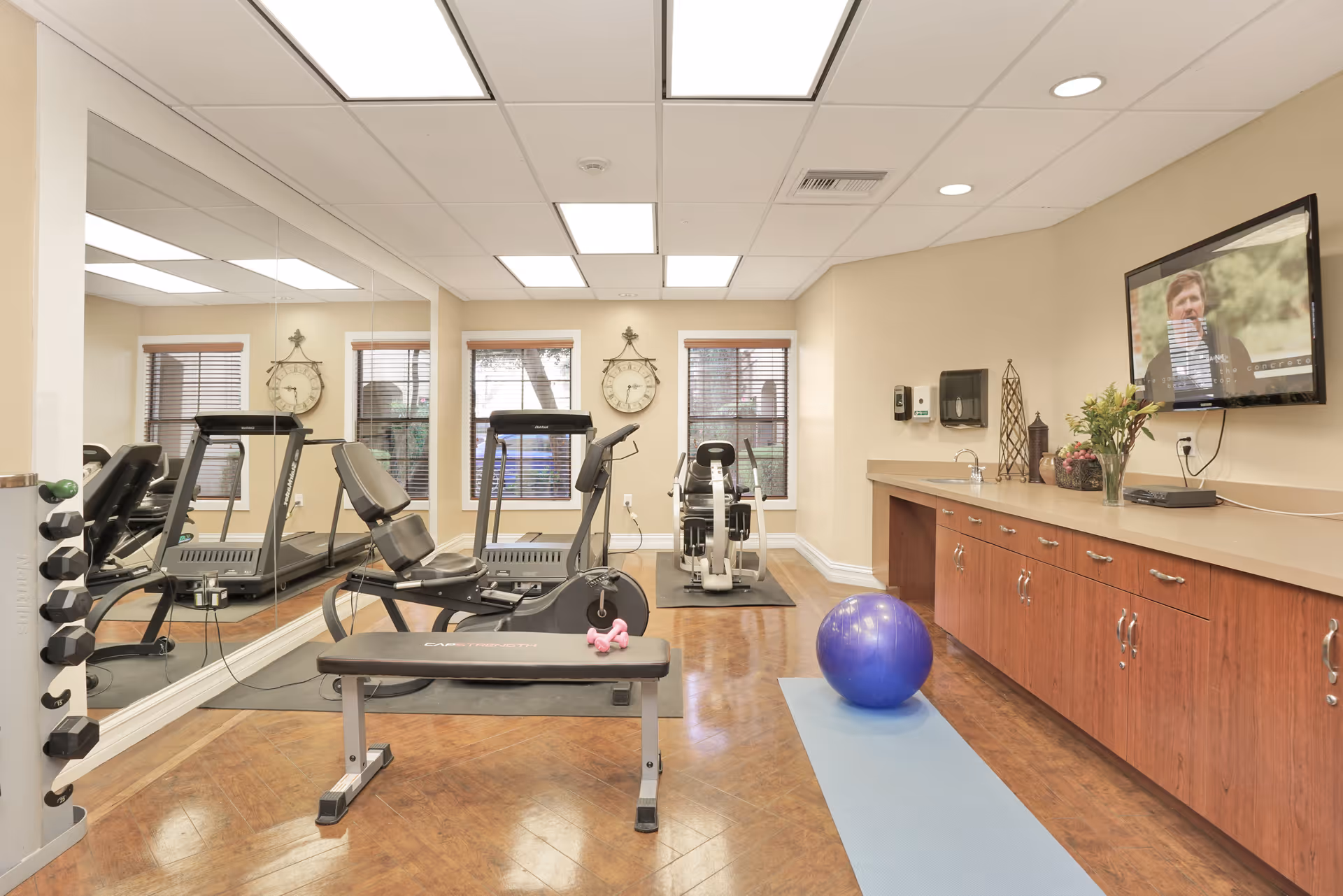 A small fitness room with exercise equipment including a recumbent bike, rowing machine, and weight bench with dumbbells. There is a large mirror on one wall, two windows with blinds, a blue exercise mat with a purple exercise ball on the floor, and a wall-mounted TV showing a person speaking. The room has wooden flooring and beige walls with a long countertop and cabinets underneath.