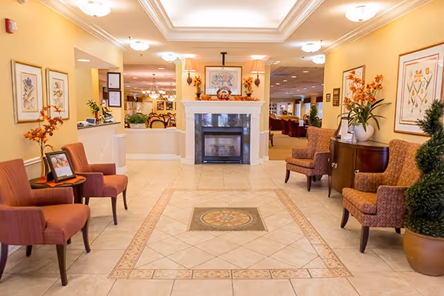 A warmly lit interior common area with beige tiled floor featuring a decorative tile pattern in the center. The room has a white fireplace with framed artwork and autumn-themed decorations on the mantel. There are upholstered chairs in shades of brown and orange arranged along the walls, with framed floral artwork hanging above. The space opens into a larger room with more seating and tables visible in the background.