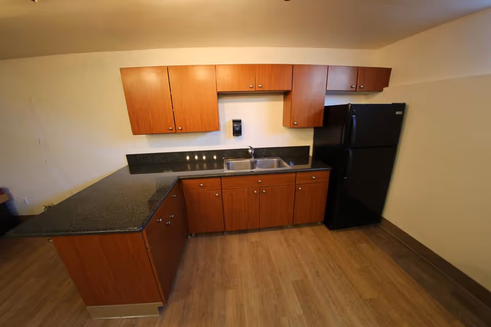 A kitchen area with wooden cabinets, a black granite countertop, a double stainless steel sink, and a black refrigerator. The floor is wood-patterned, and the walls are light-colored.