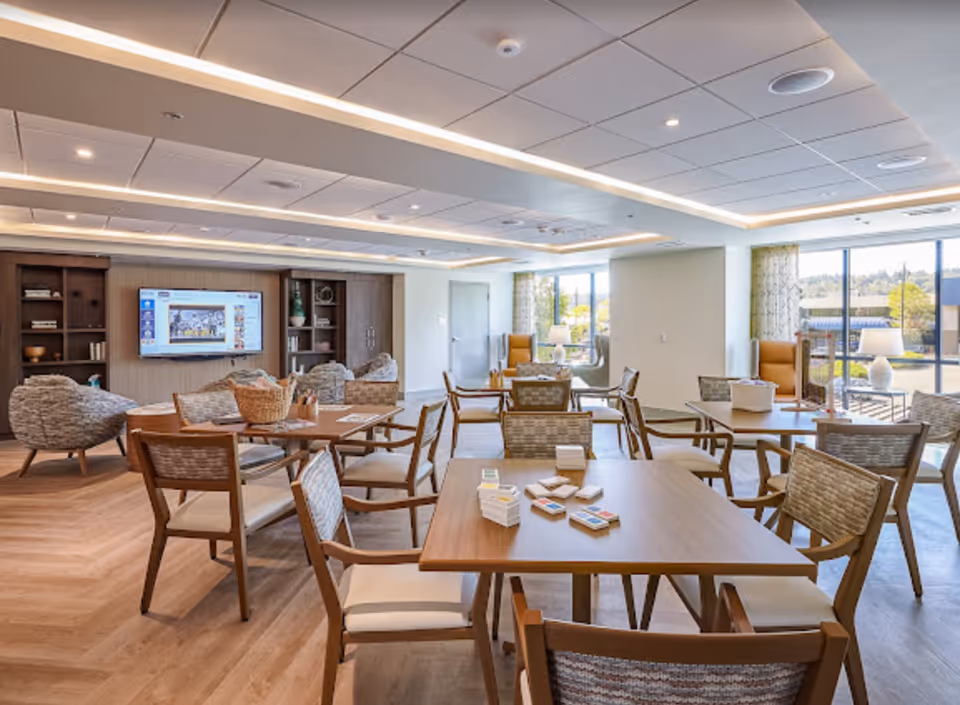 A bright and spacious common area in an assisted living facility with multiple wooden tables and chairs arranged for group activities. The room features large windows letting in natural light, a wall-mounted TV, built-in shelving with decorative items, and comfortable armchairs. The ceiling has recessed lighting and modern design elements.