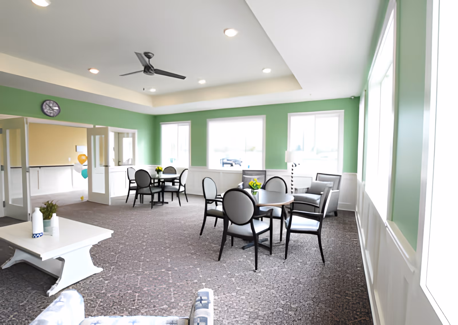 Bright common lounge and dining area with round tables, chairs, armchairs, green walls and large windows.