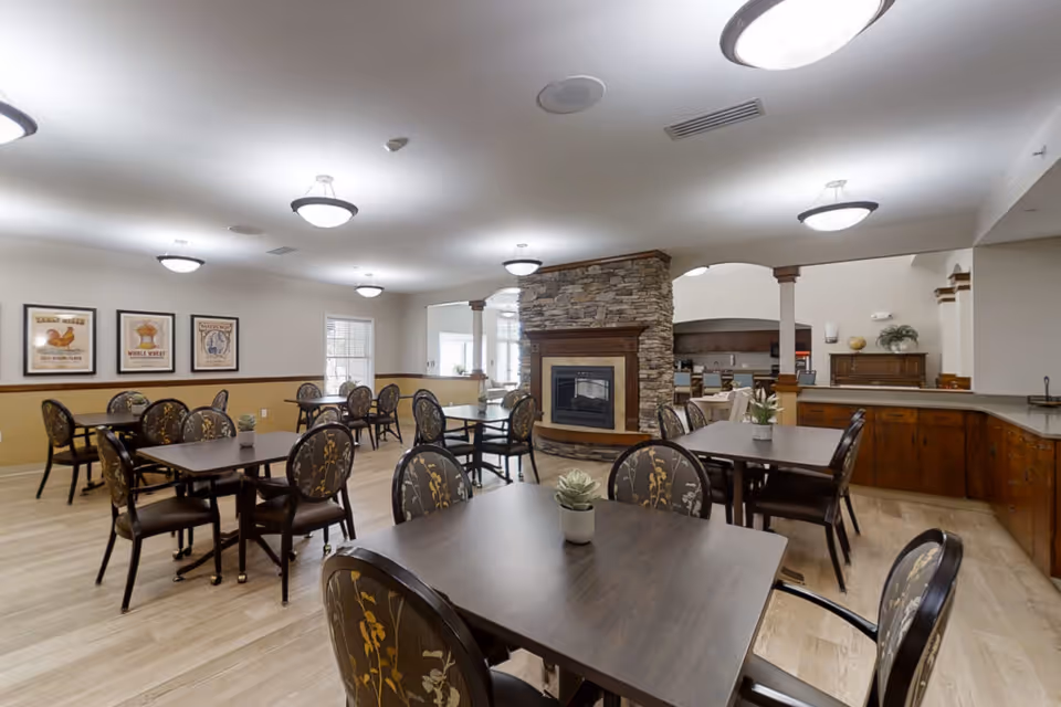 A spacious dining room with multiple dark wood tables and floral-patterned chairs arranged neatly. The room features a stone fireplace in the center, light wood flooring, and framed artwork on the walls. There are ceiling lights providing bright illumination and a counter area with wooden cabinets in the background.