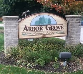 A wooden sign for Arbor Grove Assisted Living & Memory Care mounted between two stone pillars, surrounded by green plants and grass with trees in the background.
