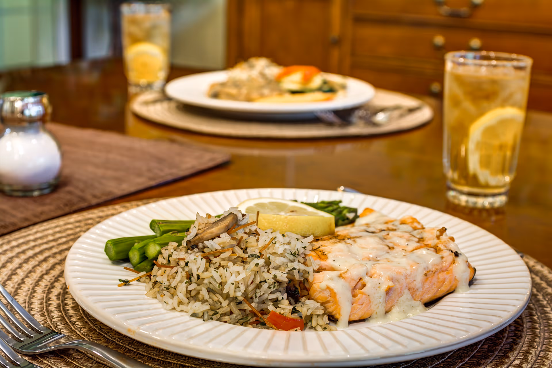 A close-up of a plated meal featuring a piece of salmon topped with a creamy white sauce, a serving of seasoned rice with vegetables, and steamed asparagus, accompanied by a glass of iced tea with a lemon slice on a wooden table set with placemats and silverware.