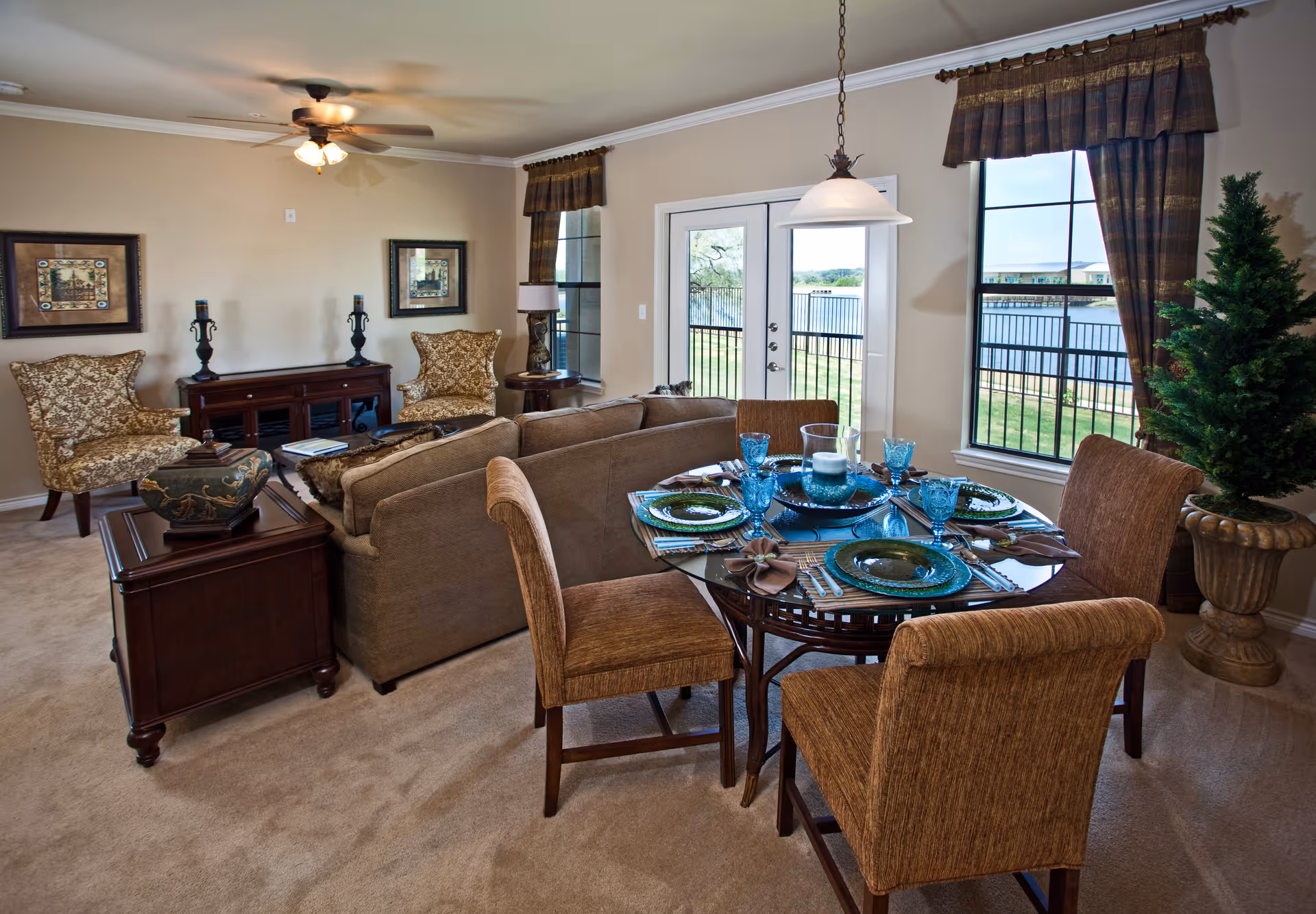 A cozy living and dining area in a senior living facility featuring a round glass dining table set with blue glassware and green plates, surrounded by four upholstered chairs. Behind the dining area is a beige sofa facing a wooden console table with decorative items and two patterned armchairs. The room has beige walls, carpeted floor, two large windows with brown curtains, and a glass door leading outside with a view of a fenced grassy area and water beyond.