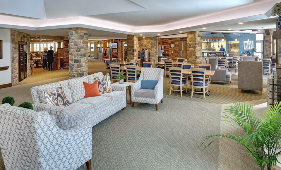 Spacious and well-lit common area in a retirement community featuring patterned sofas and armchairs, multiple wooden dining tables with chairs, stone pillars, and a kitchen area in the background. Two people are seen conversing near the entrance to another room.