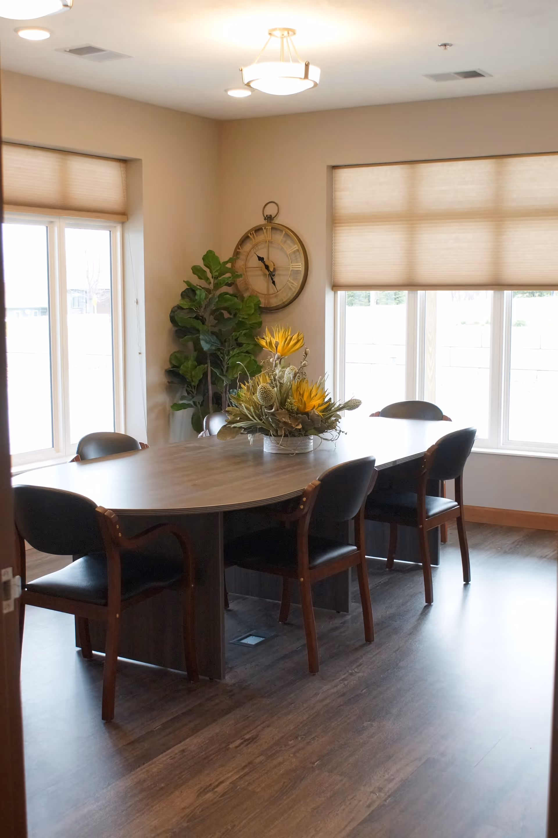 A dining room with a large oval wooden table surrounded by six black cushioned chairs. A decorative floral arrangement with yellow flowers is placed in the center of the table. The room has large windows with beige blinds, a wall clock, and a tall green plant in the corner. The floor is wooden, and the ceiling has modern light fixtures.
