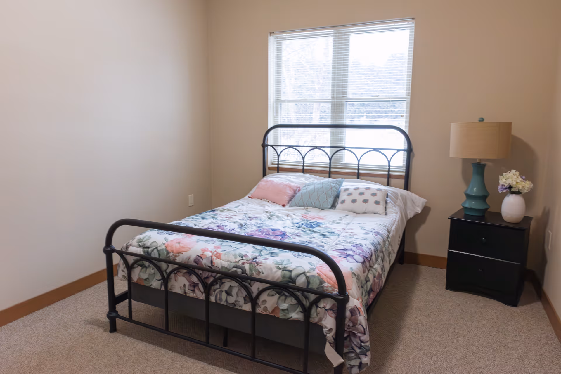 A bedroom with a black metal bed frame, floral patterned bedding, and three decorative pillows. There is a window with blinds behind the bed, and a black nightstand to the right of the bed with a teal lamp and a white vase with flowers.