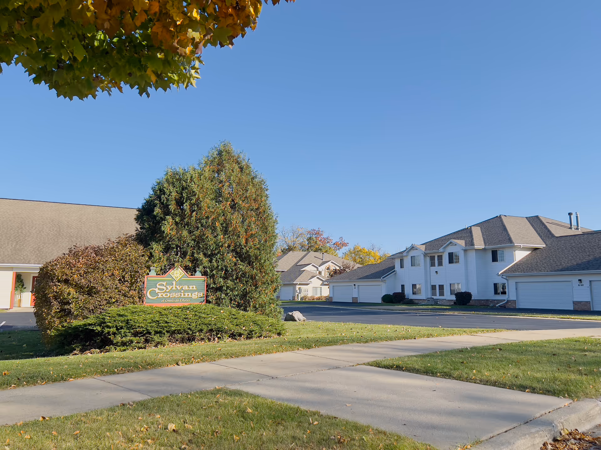 Exterior view of a low-rise senior living complex with white buildings, garages, landscaping, and a sign reading "Sylvan Crossings".