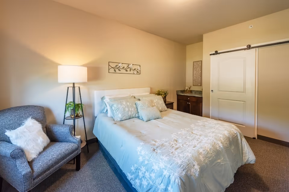 Furnished bedroom with a bed dressed in light blue floral bedding, an upholstered armchair, floor lamp, and a sliding barn door.