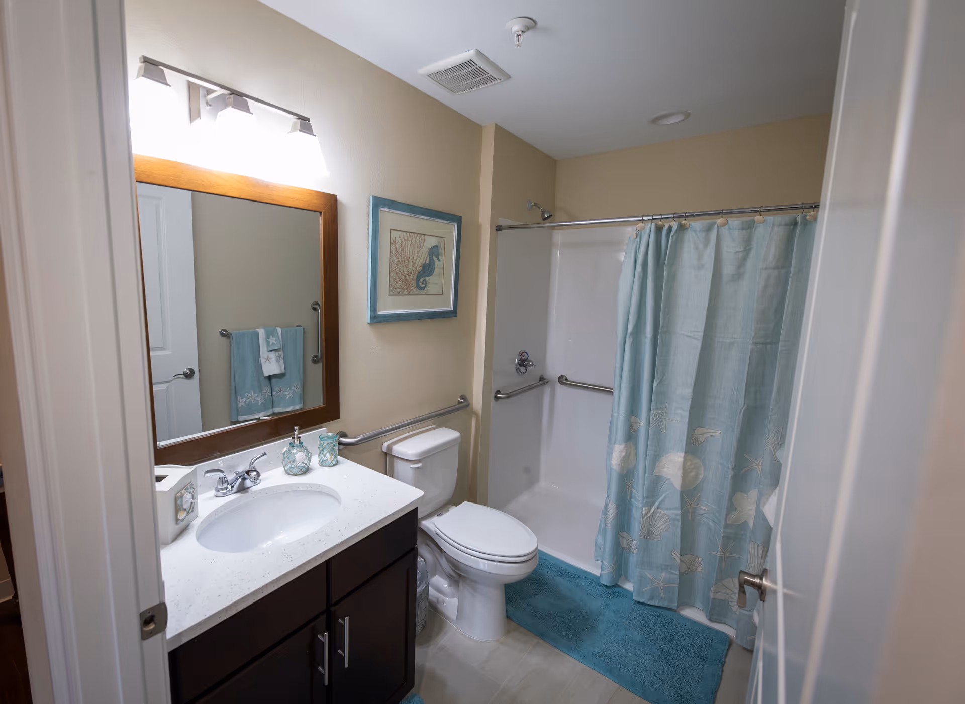 A bathroom with a white sink and countertop, dark wood vanity, large mirror with three lights above it, toilet, and a shower with a blue curtain featuring seashell and starfish designs. There is a blue bath mat on the floor and a framed picture of a seahorse on the wall.