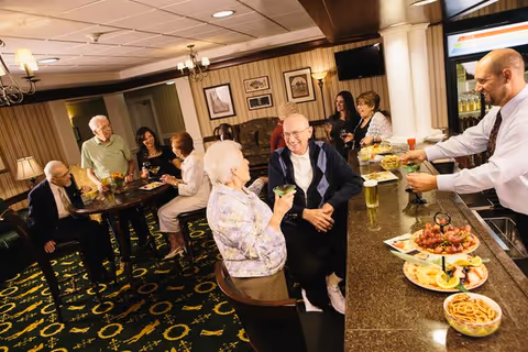 A group of elderly people socializing and enjoying drinks and snacks in a warmly lit lounge area with a bar counter. A bartender is serving drinks while others are seated or standing around tables, engaging in conversation and smiling.