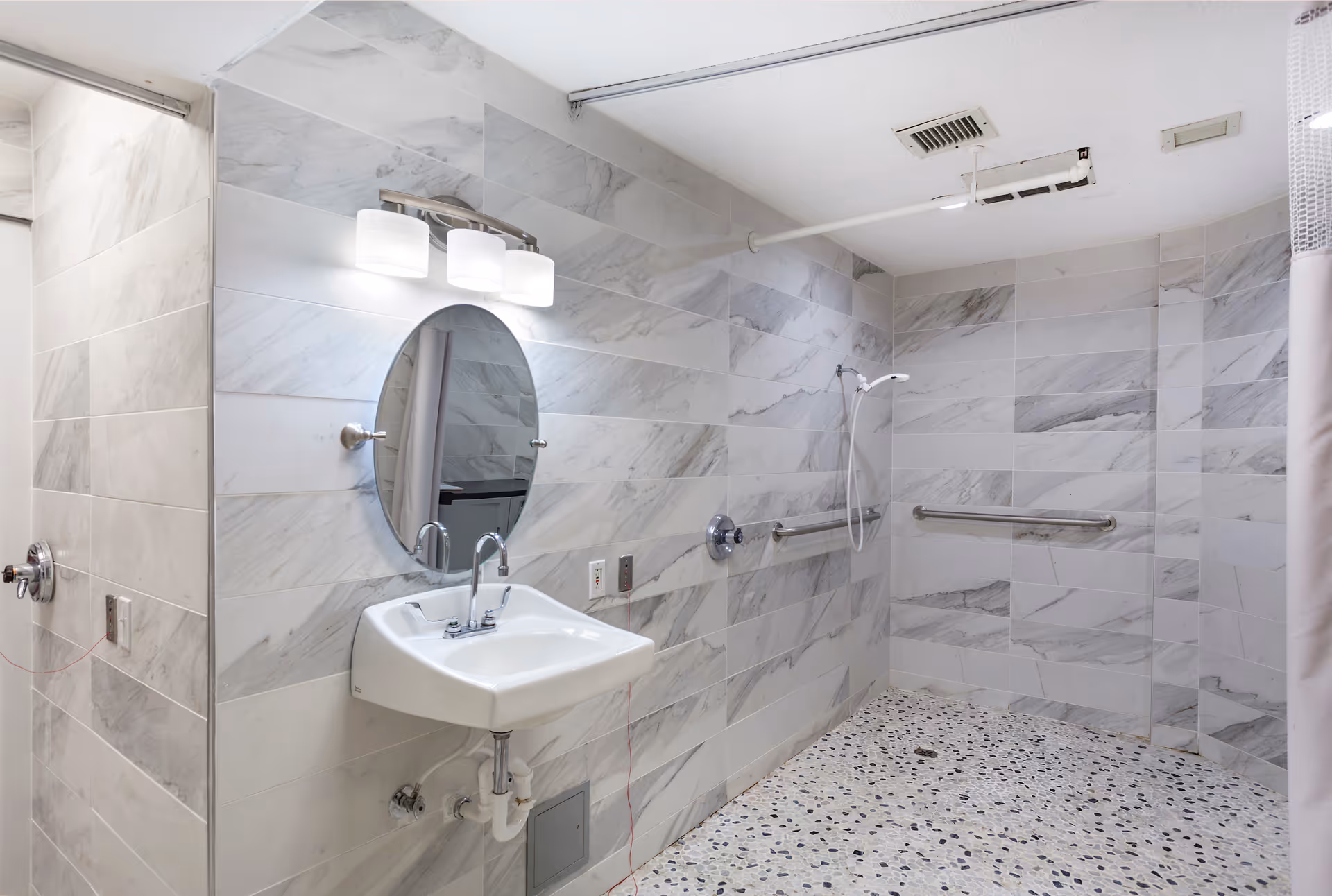 Accessible bathroom with a wall-mounted sink beneath an oval mirror, handheld shower, grab bars, and tiled walls with a pebble-pattern floor.
