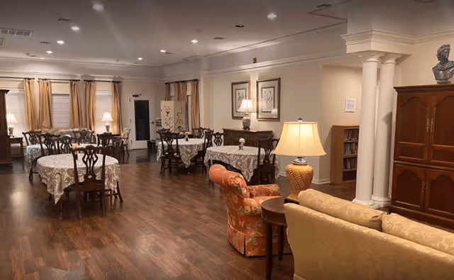 Interior view of a senior living facility dining room with multiple round tables covered with white lace tablecloths and wooden chairs. The room has hardwood flooring, beige walls, and several lamps providing warm lighting. There are curtains on the windows and framed artwork on the walls. In the foreground, there is a seating area with a floral armchair, a beige sofa, and a side table with a lamp.