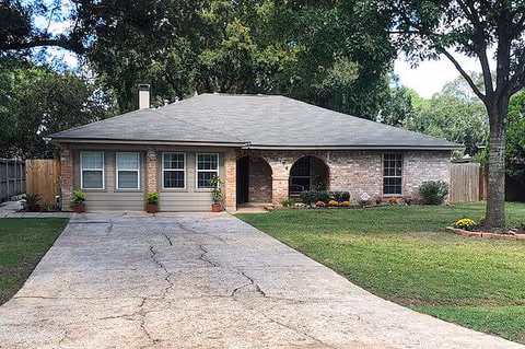 Single-story brick and siding house with a gray roof, a concrete driveway, and a well-maintained front yard with grass, trees, and small flower beds.