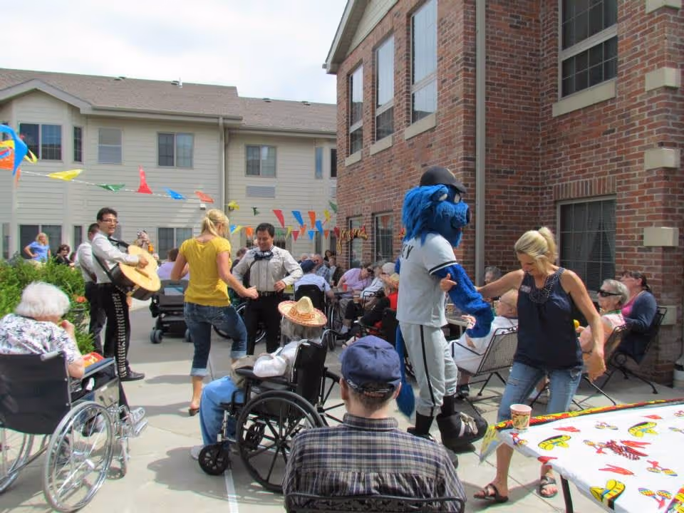 Outdoor courtyard at a senior living facility with elderly residents sitting in wheelchairs and chairs, people dancing, a person in a blue mascot costume, and musicians playing guitars. Colorful pennant banners are strung overhead between buildings.