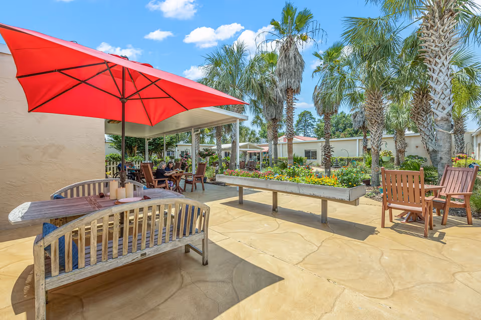 Outdoor courtyard with patio seating under a red umbrella, palm trees, and raised flower planters.