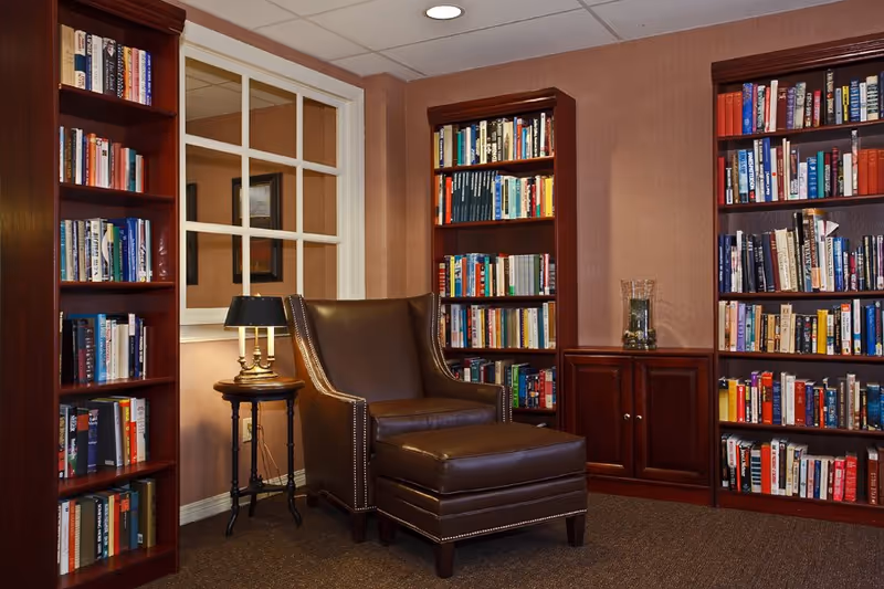 A cozy reading nook in a library or common area featuring a dark brown leather armchair with a matching ottoman, surrounded by wooden bookshelves filled with books. A small round side table with a lamp is positioned next to the chair, and a window with white framing is visible on the wall behind.