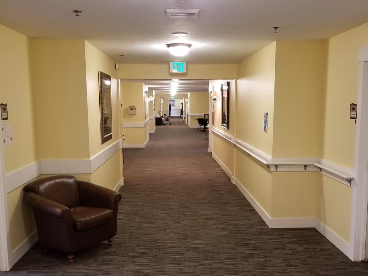 A long, well-lit hallway in an assisted living facility with yellow walls, brown carpet, handrails on both sides, and a brown leather armchair on the left. There are framed pictures on the walls and an exit sign at the far end. Some chairs and a person with a walker are visible in the distance.