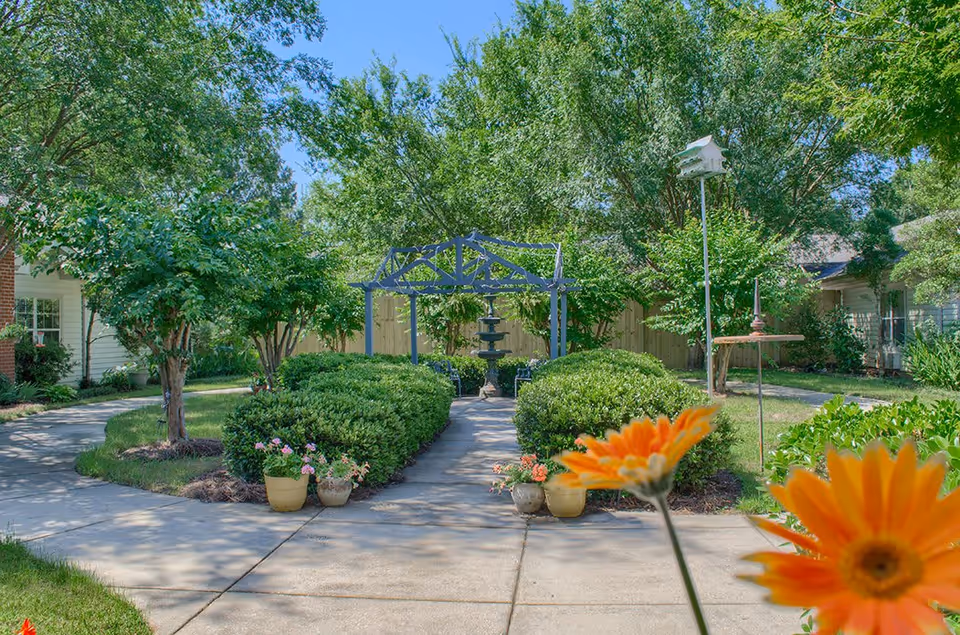 A peaceful outdoor garden area with a paved walkway leading to a blue pergola and a tiered water fountain surrounded by green bushes and trees. There are potted flowers along the walkway and a birdhouse on a tall pole to the right. Bright orange flowers are visible in the foreground.