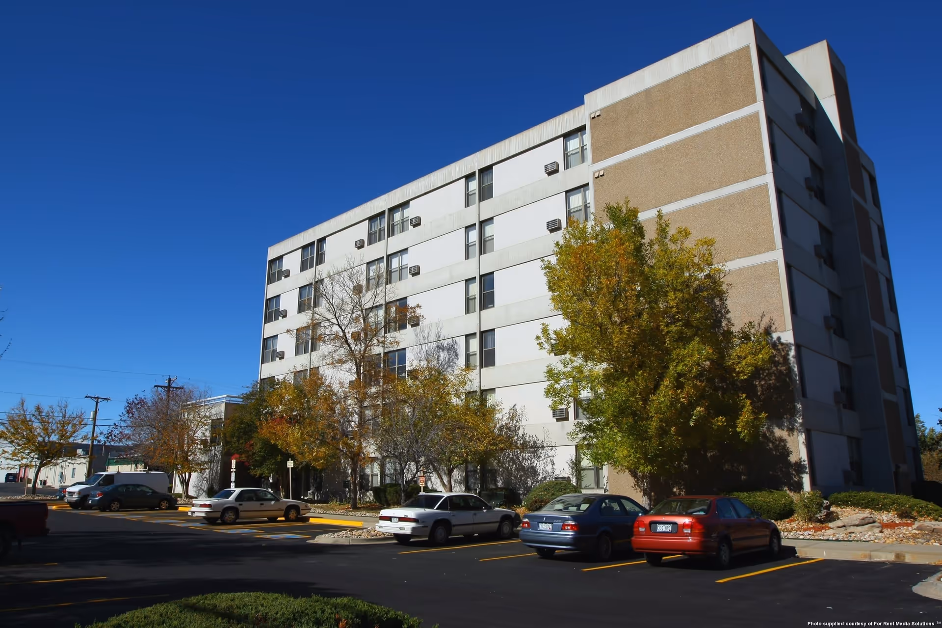 Six-story apartment building with trees and a parking lot of cars under a clear blue sky.