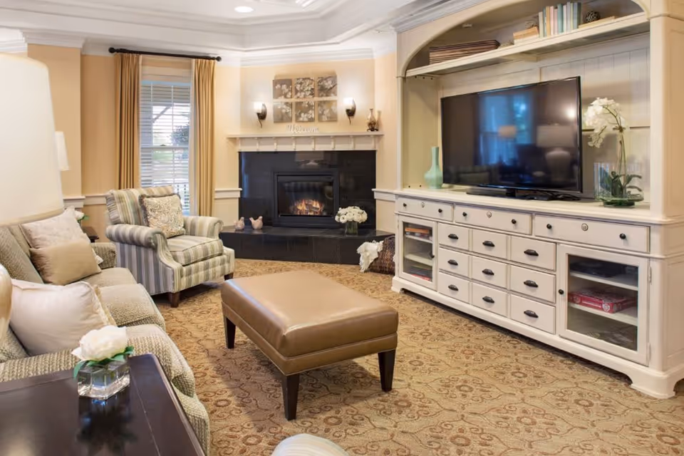 A cozy living room with a beige patterned carpet, a striped armchair, a beige sofa with multiple cushions, and a brown leather ottoman in the center. There is a white entertainment unit with a flat-screen TV and decorative items including a vase with flowers. A fireplace with a black surround is in the corner, above which are wall decorations and a 'Welcome' sign. The room has beige walls, a window with beige curtains, and soft lighting.
