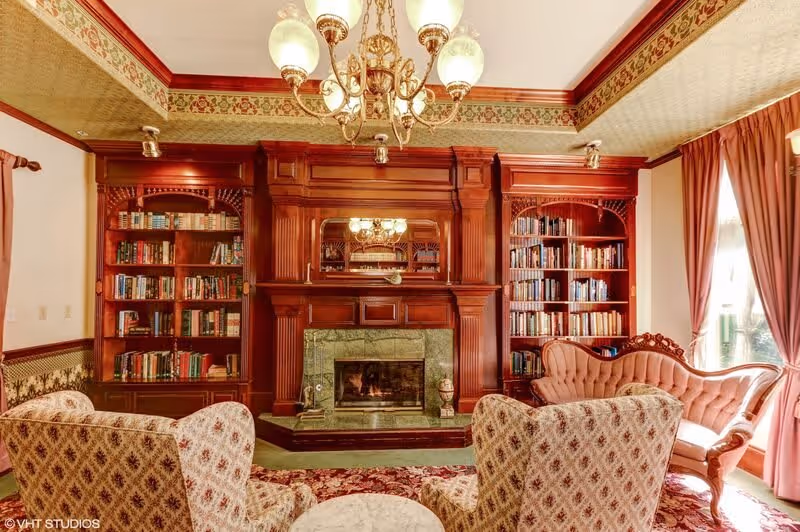 A cozy living room with vintage decor featuring a large wooden fireplace with a green marble surround, flanked by built-in bookshelves filled with books. The room has patterned wallpaper and a decorative border near the ceiling. There are two upholstered armchairs with a floral pattern and a pink tufted sofa. A chandelier with multiple glass globes hangs from the ceiling, and large windows with pink curtains allow natural light to enter.