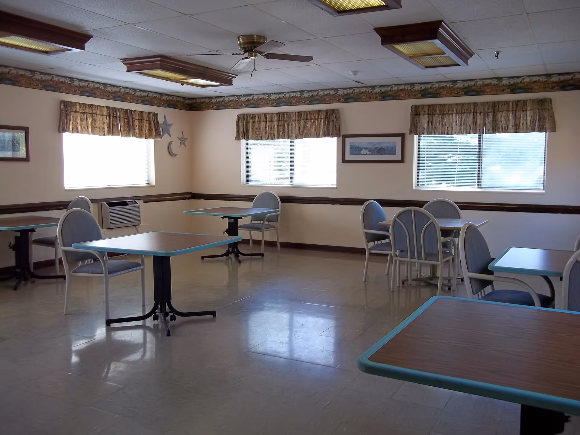 A dining room with several tables and chairs arranged around the room. The walls have windows with valance curtains, framed pictures, and decorative stars and a crescent moon. The ceiling has fluorescent lights and a ceiling fan. The floor is tiled and the room appears clean and well-lit.