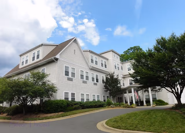 Exterior view of a large, multi-story senior living facility building with white siding, numerous windows, and a covered entrance. The building is surrounded by well-maintained landscaping including trees, bushes, and a curved driveway. The sky is partly cloudy with patches of blue.