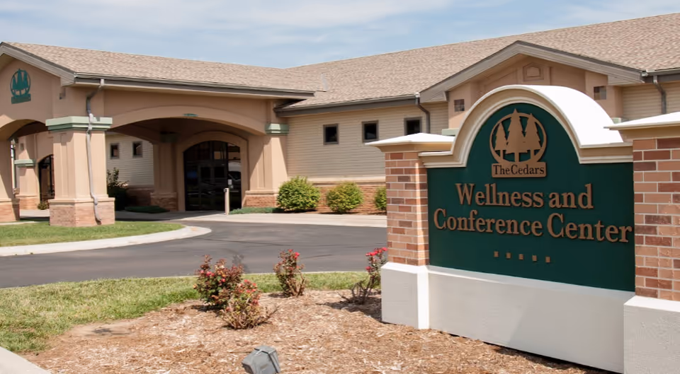 Exterior view of The Cedars Wellness and Conference Center building with a sign in the foreground displaying the facility name. The building has a beige facade with a covered entrance and some landscaping with bushes and flowers.