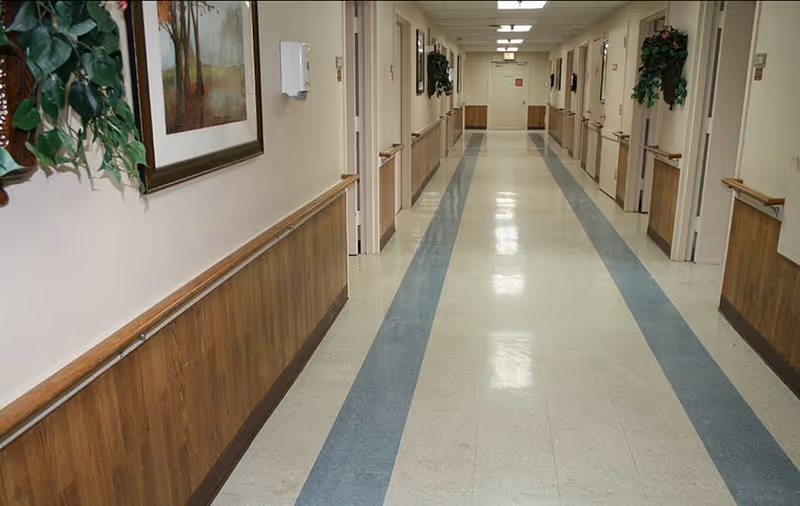 Long empty interior hallway in a rehabilitation center with handrails, doors on both sides, framed artwork and hanging plants.