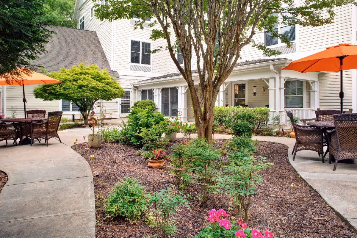 Outdoor courtyard area of a senior living facility with a garden bed containing various shrubs and flowers, surrounded by a concrete walkway. There are two seating areas with brown wicker chairs and tables, each shaded by an orange umbrella. The building exterior is white with multiple windows and a covered porch area.