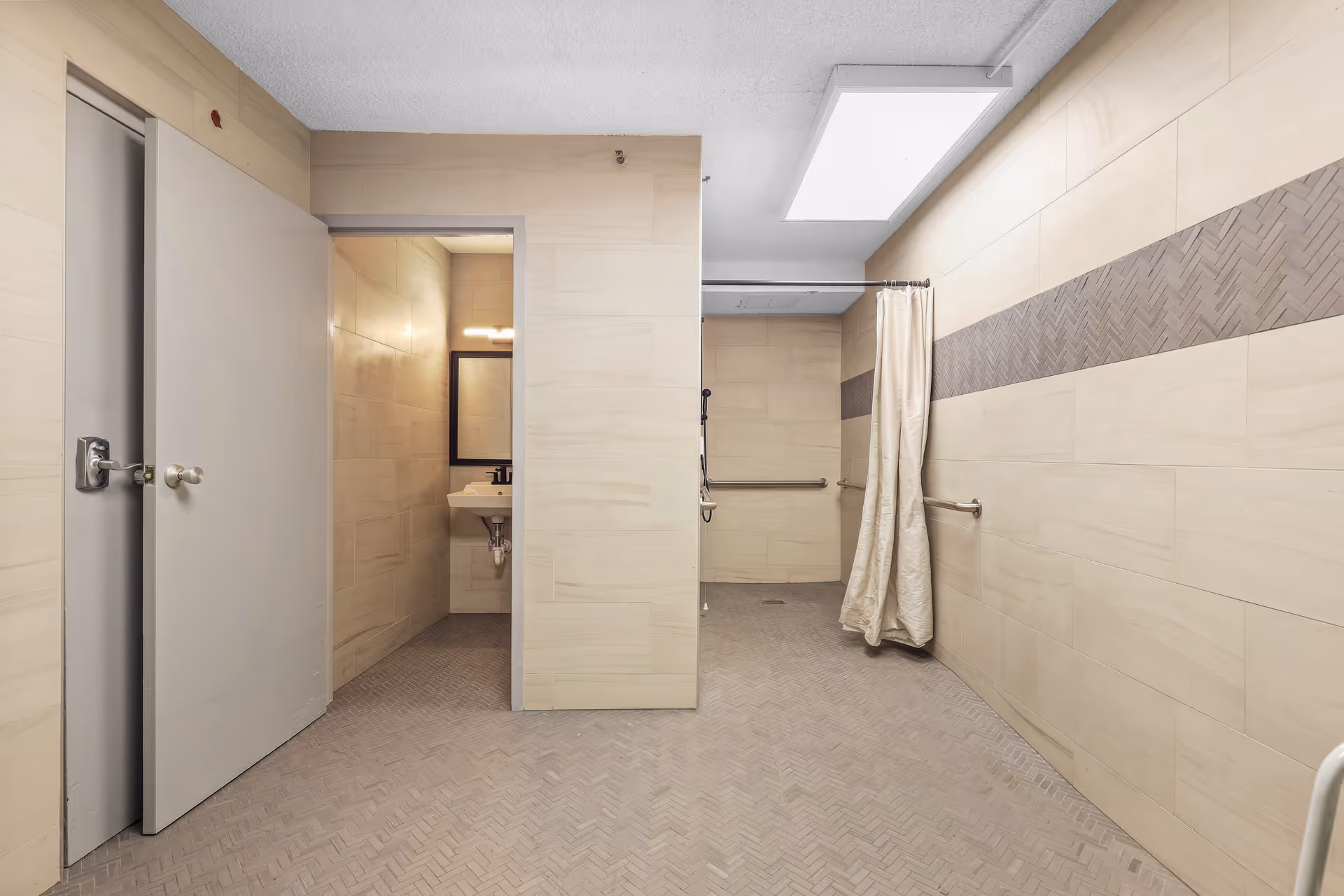 A spacious, accessible bathroom with beige tiled walls and floor. The room features a shower area with a curtain and grab bars, a sink with a mirror and light above it, and two doors, one partially open. The lighting is bright with a large ceiling light panel.